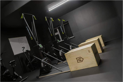 Gym equipment including rowing machines and wooden boxes in a dark room.